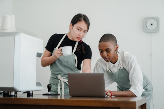 Serious Asian female barista with coffee in hand teaching new black colleague to use computer program while standing at table with laptop
